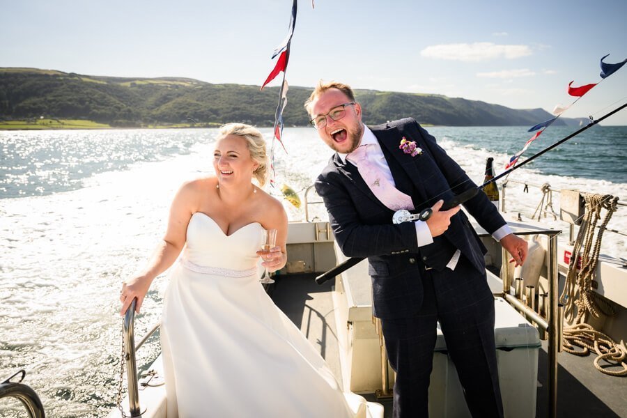 Bride and groom on a boa in porlock weir. Both very happy and the groom is holding a fishing rod.
